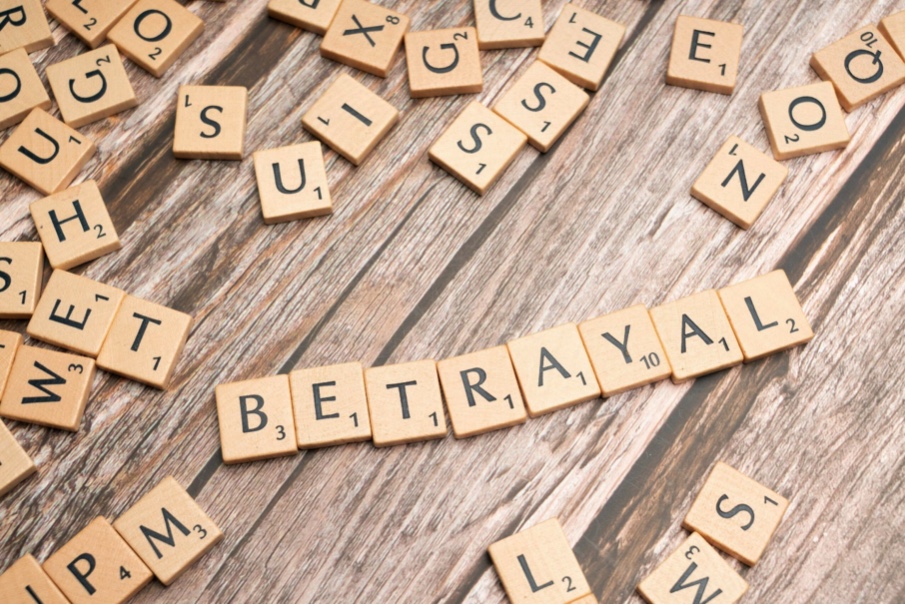 Ułożone z drewnianych kostek słowo 'Betrayal' – zdrada – na tle liter i drewnianych desek; emocjonalny motyw poruszenia i straty.